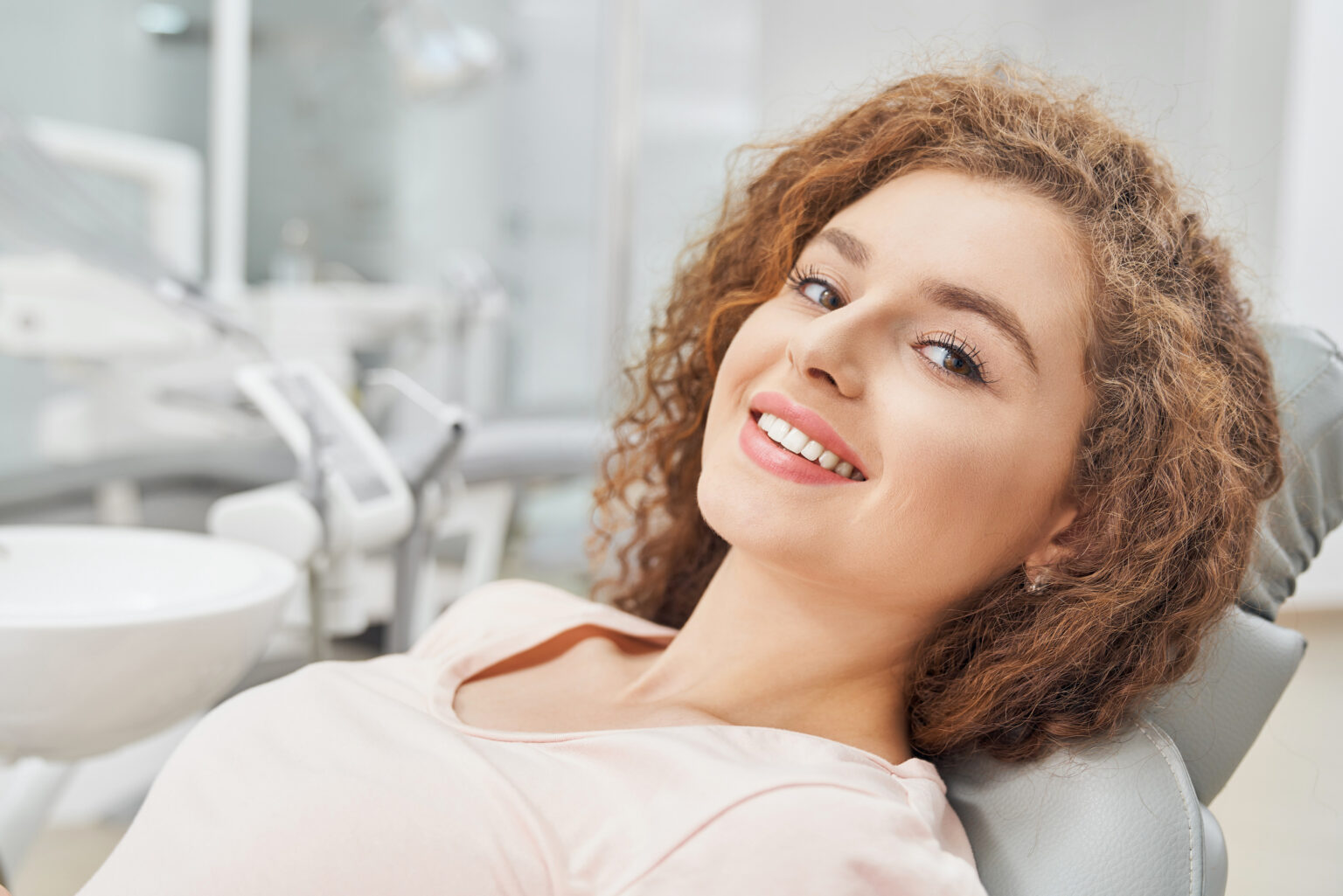 Beautiful female patient with curly hair in pink shirt sitting in dental clinic and waiting for teeth examination, smiling and looking at camera. Concept of medicine and treatment